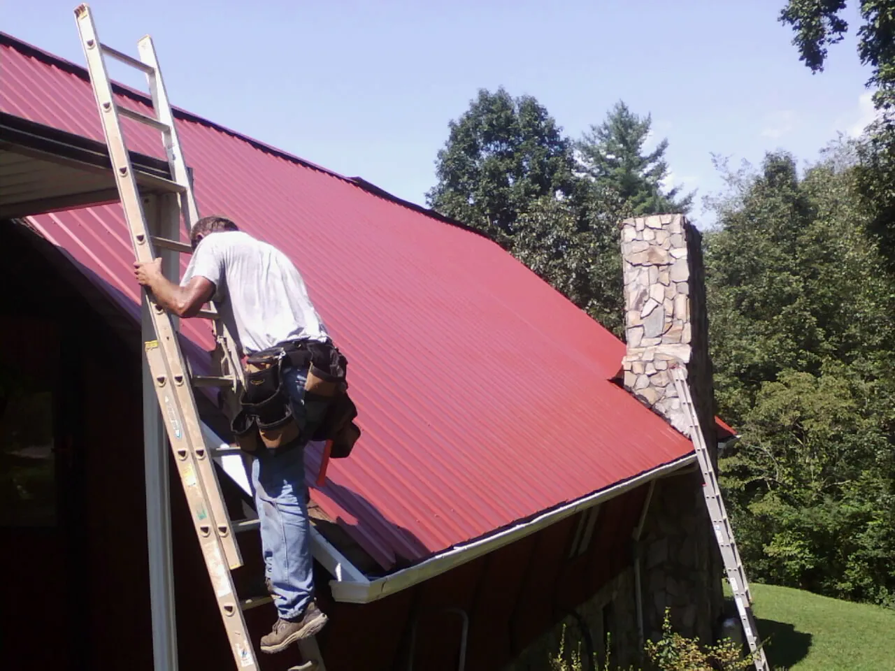 Roofer on a ladder beside a red metal roof and stone chimney — Soffit & Fascia Repair by Roofing Brothers in McMinnville